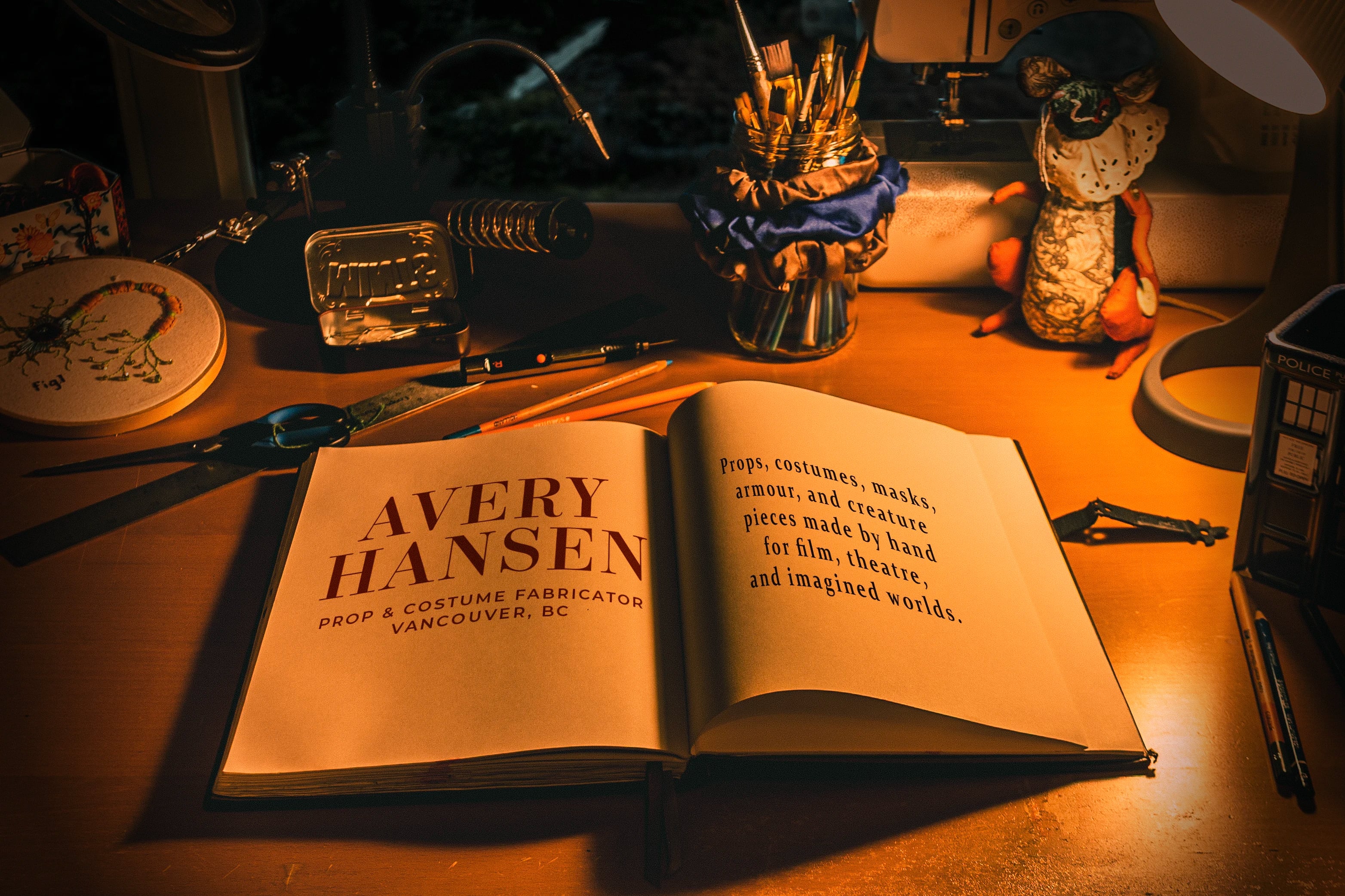Avery Hansen's lamp-lit workbench at night: an open book reading 'Avery Hansen, Prop & Costume Fabricator, Vancouver BC' and 'Built for Story: props, costumes, masks, armour and creature pieces made by hand for film, theatre, and imagined worlds', surrounded by a sewing machine, magnifier lamp, brushes, and handmade oddities.