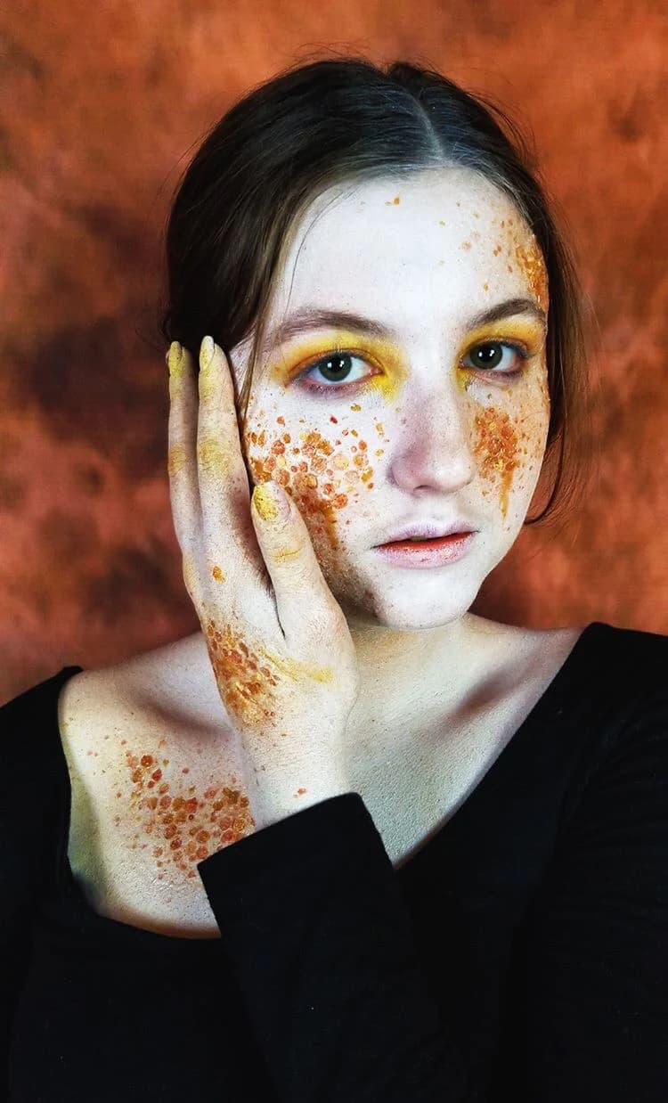 SFX makeup portrait: a performer with amber honeycomb-textured prosthetic transfers across one cheek, hand, and chest, against a rust-coloured backdrop.