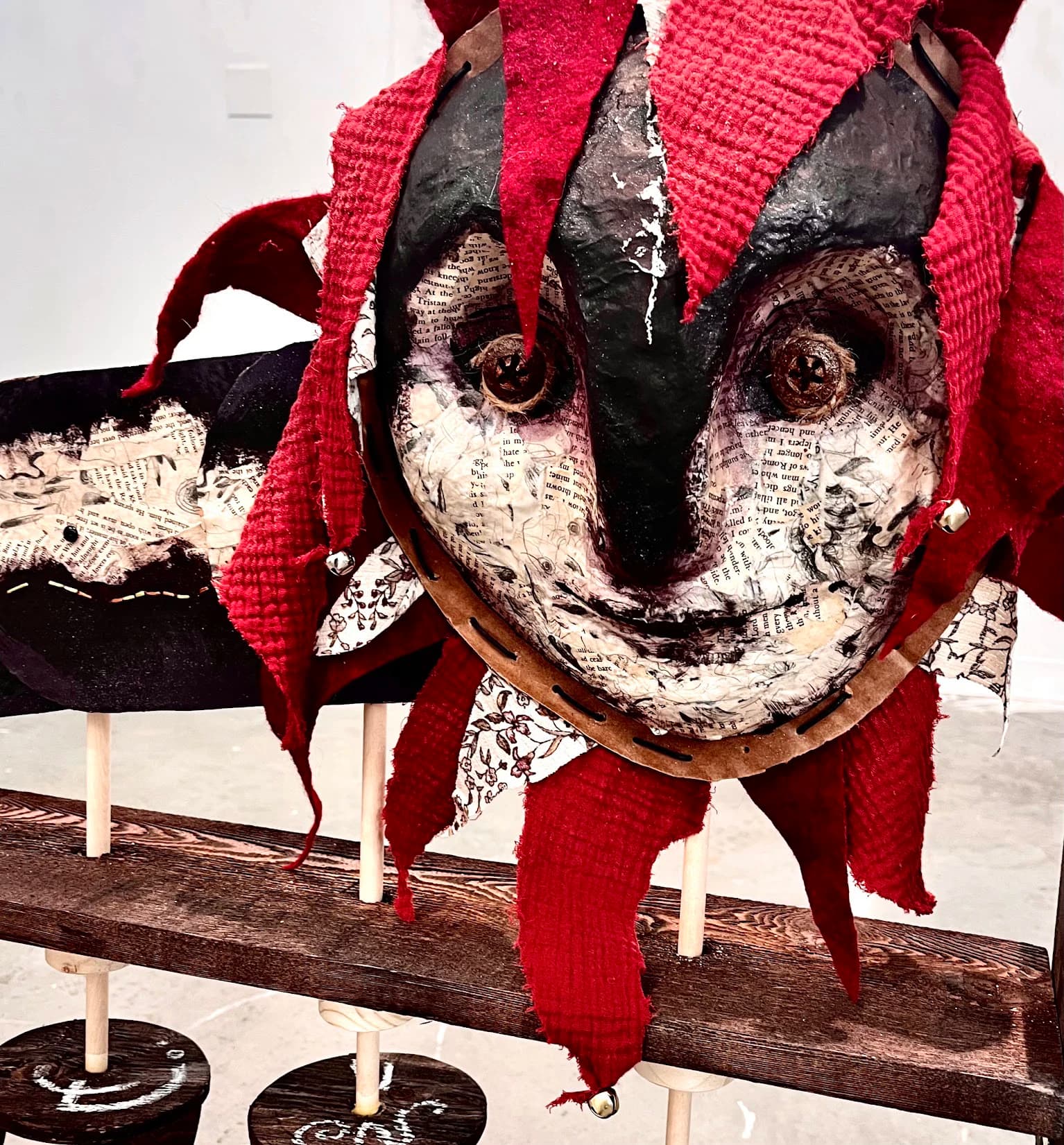 Close-up of the serpent's papier-mâché face: black-and-cream book-page skin, button-like eyes, and torn red fabric fringe hung with small bells.