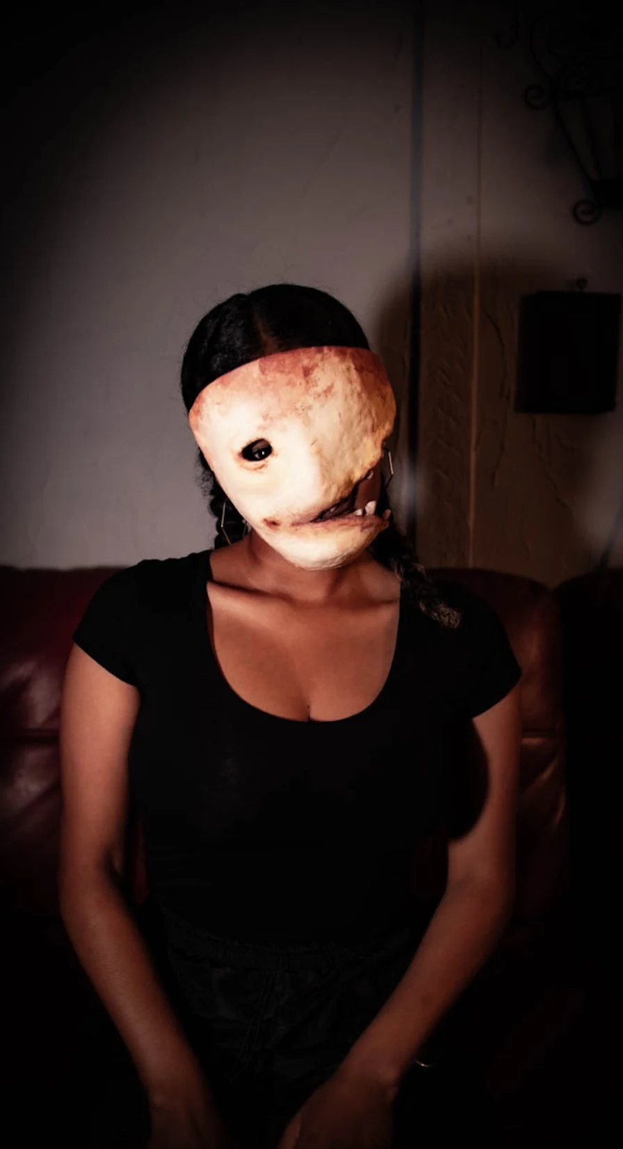 Seated performer in black wearing the One-Eye mask: a rounded pale face with a single dark eye and a crooked toothy mouth, spotlit against a plaster wall.