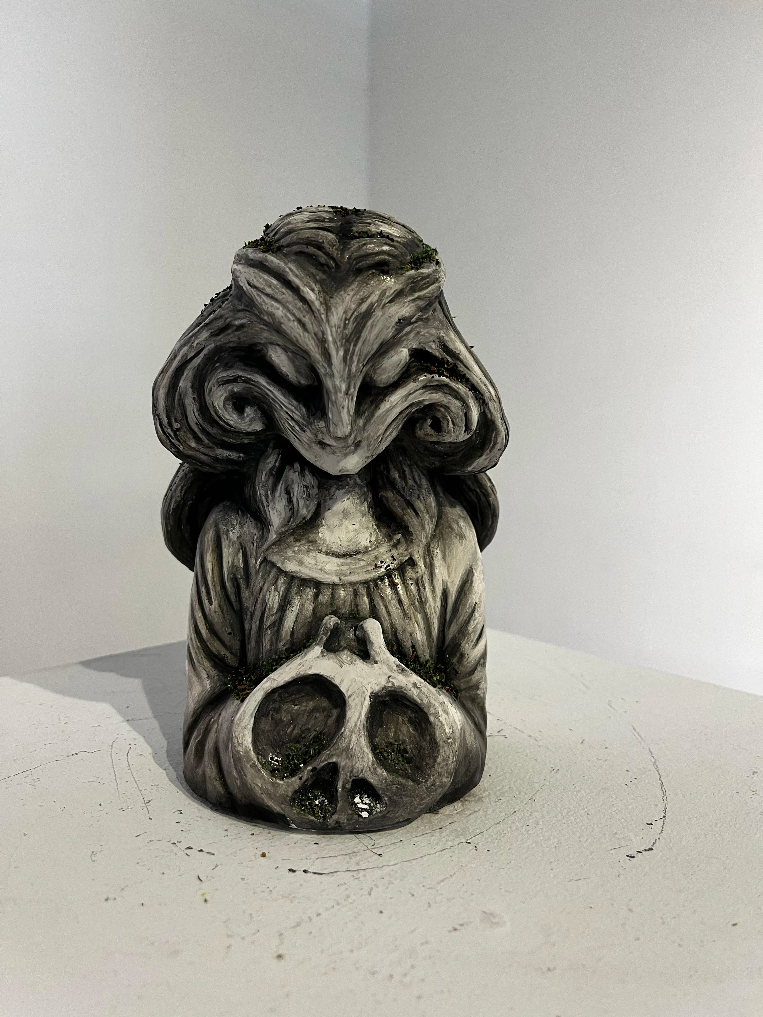 Front view of a small cast concrete statuette: a hooded creature with swirling carved hair holding a skull, with moss growing in its crevices, on a white plinth.